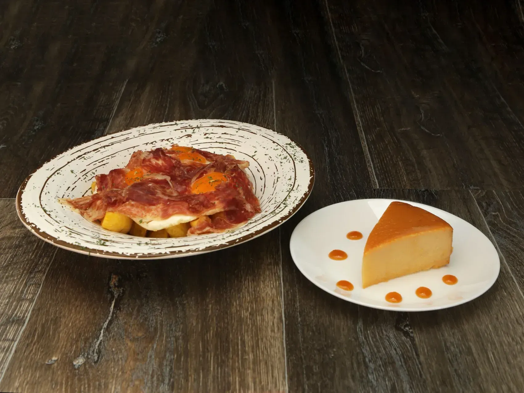 Plato de comida con jamón y huevos, junto a un plato con flan de caramelo sobre una mesa de madera oscura.