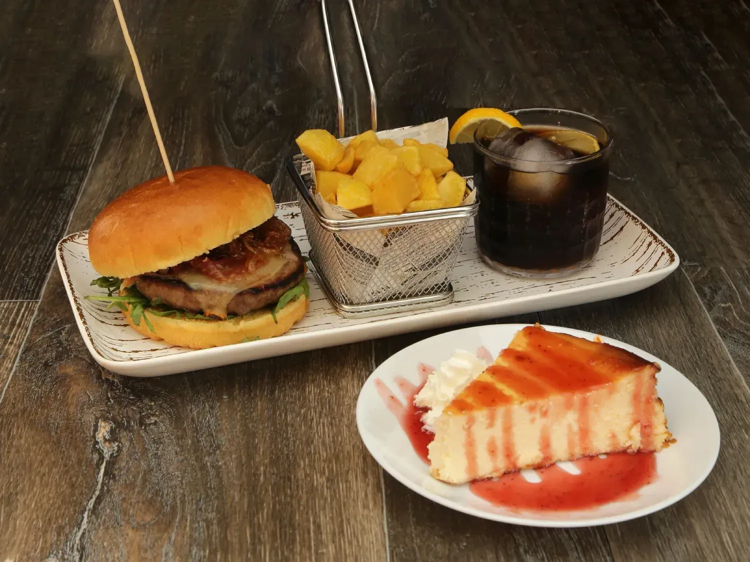 Hamburguesa, patatas fritas, bebida y tarta de queso en una bandeja, dispuesta sobre una mesa de madera.