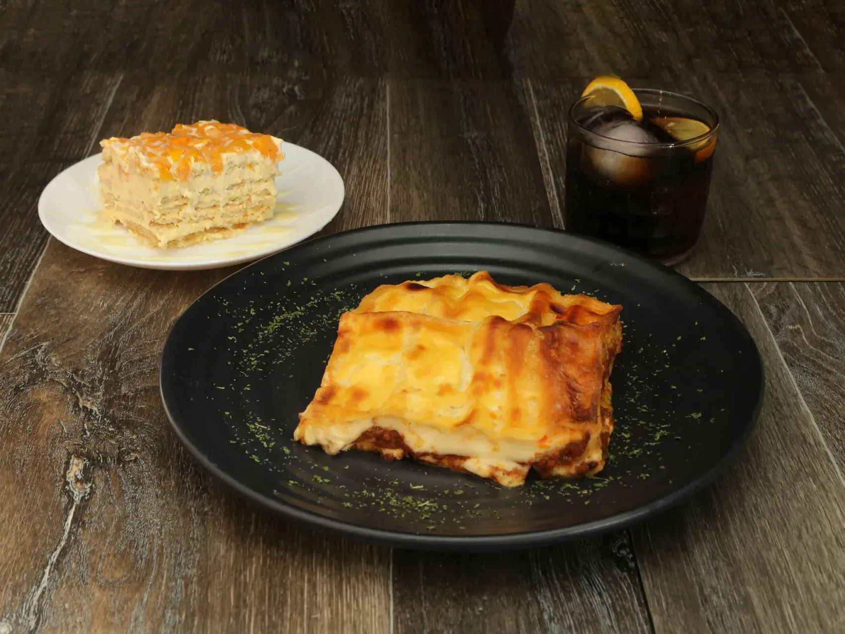 Lasaña en un plato negro, pastel y una bebida con limón sobre una mesa de madera.