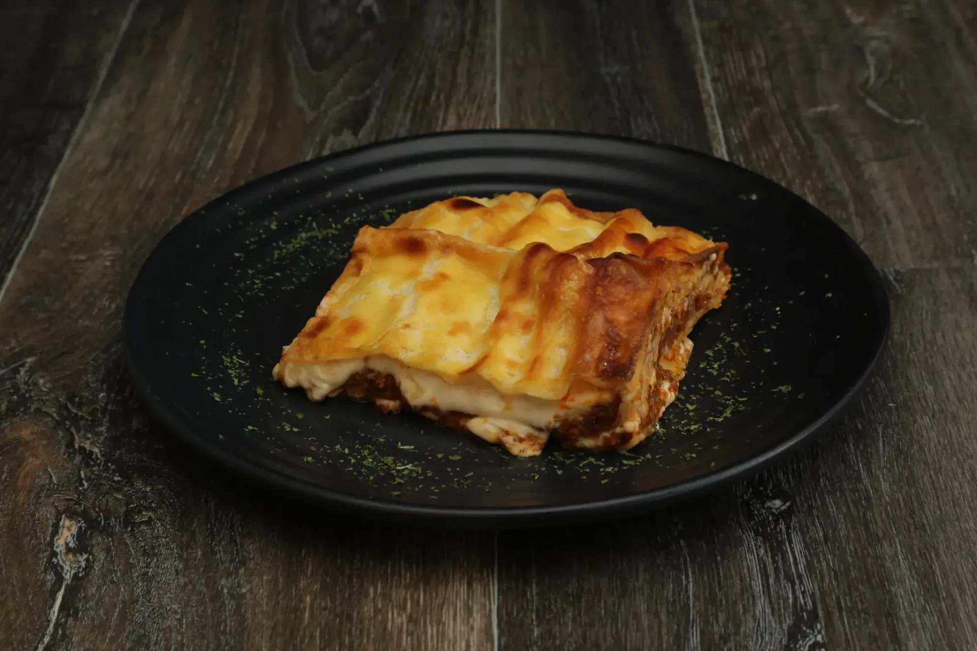 Lasaña en un plato negro, sobre una superficie de madera oscura.