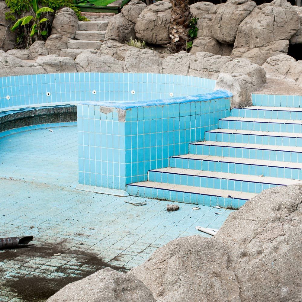 Piscina vacía, revestida de azulejos azules, rodeada de rocas y escalones.