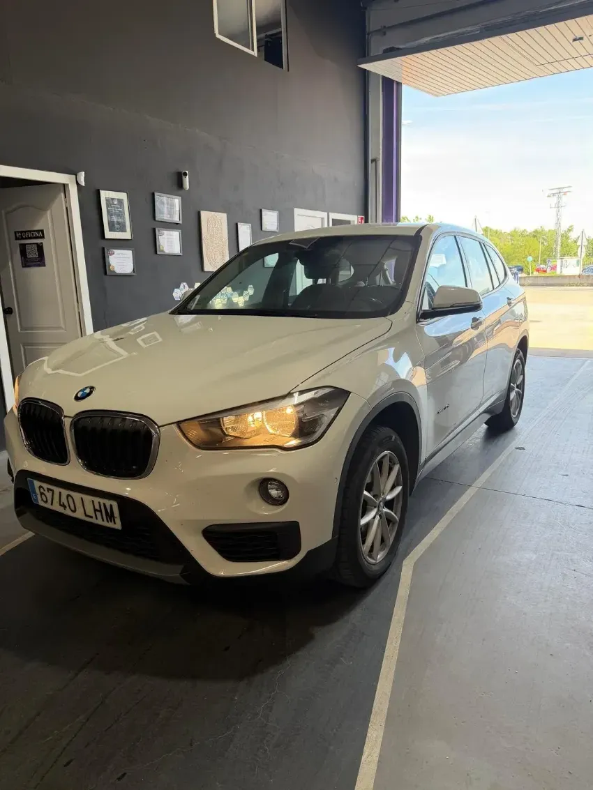 SUV BMW blanco estacionado en la entrada de un garaje, vista frontal izquierda, con las luces encendidas.