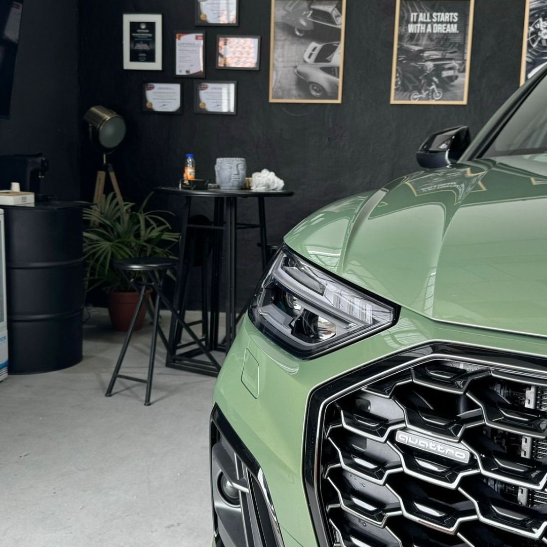 Parte delantera de un SUV verde menta en una sala de exposición oscura con obras de arte en las paredes y plantas en macetas.