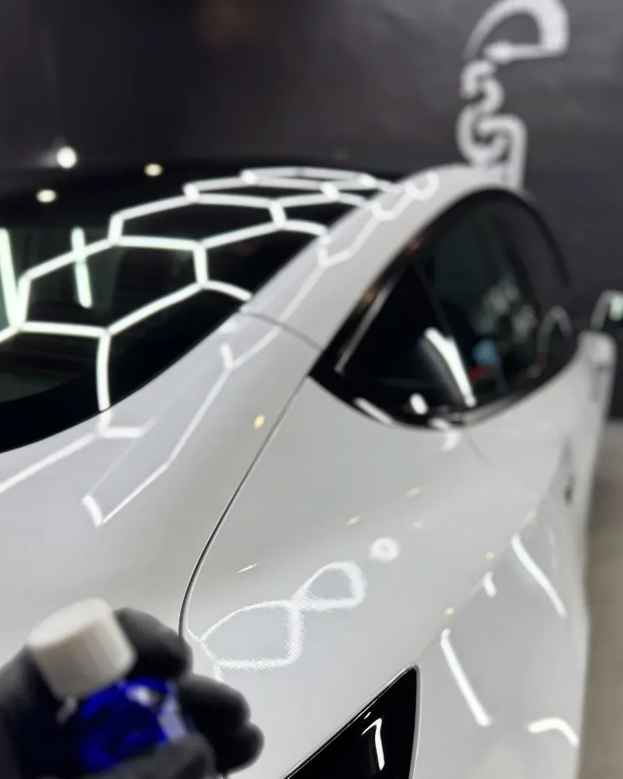 Un coche deportivo blanco en una sala de exposición, con las luces del techo y una pared estampada que reflejan la luz.