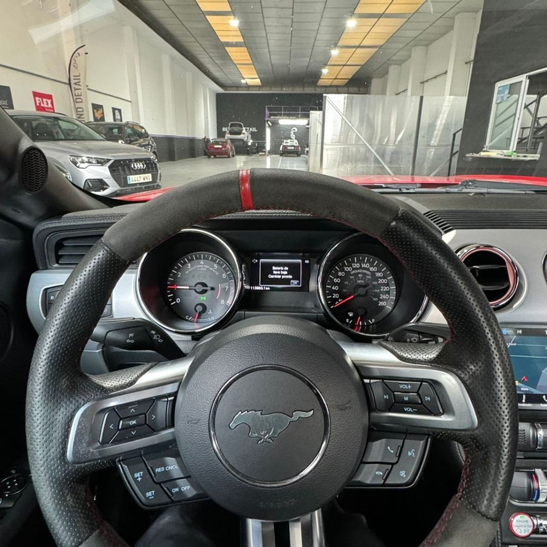 Vista desde el punto de vista del conductor del volante y el salpicadero de un Ford Mustang dentro de un garaje, con otro coche delante.