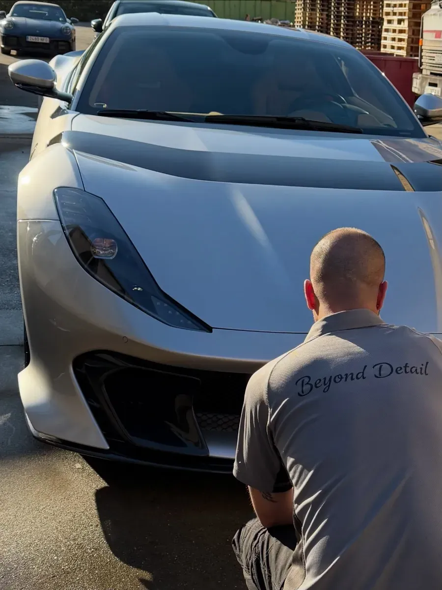 Una persona se arrodilla frente a un coche deportivo blanco en un aparcamiento al atardecer.