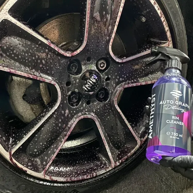 Una mano enguantada sostiene un limpiador de llantas de color morado junto a una llanta de aleación negra sucia.
