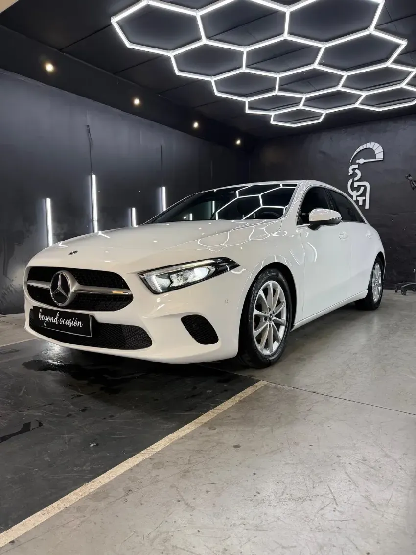 Sedán Mercedes blanco en una elegante sala de exposición con luces de techo hexagonales.