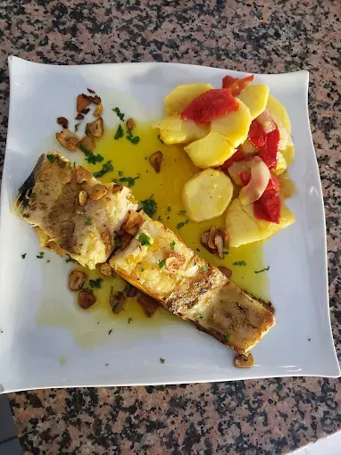 Filete de pescado a la parrilla con patatas y tomates, rociado con aceite de oliva, en un plato blanco.