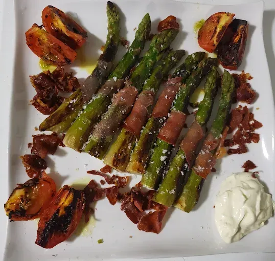 Espárragos a la parrilla envueltos en prosciutto, tomates asados y alioli en un plato blanco.