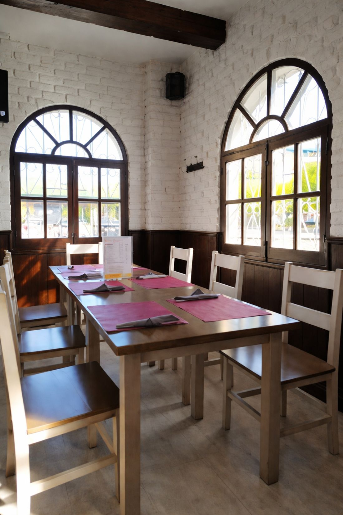 Comedor con mesa larga de madera, sillas, ventanas arqueadas y pared de ladrillo.