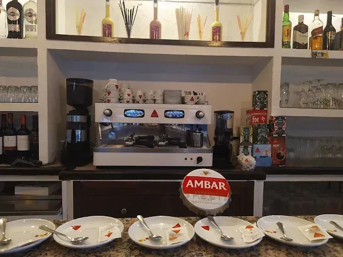Interior de bar con máquina de café expreso, botellas, vasos y platos en el mostrador.