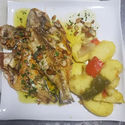 Pescado a la parrilla con patatas y pimientos en un plato blanco, rociado con aceite de oliva y perejil.