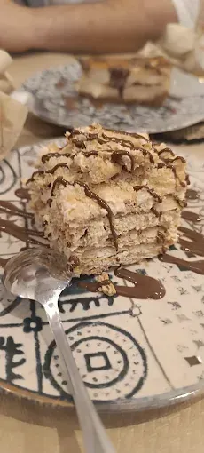 Rebanada de postre en capas rociada con chocolate sobre un plato decorativo con una cuchara.