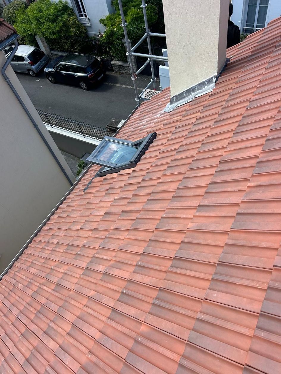 Toit en pente recouvert de tuiles rouges avec une lucarne, vu d'en haut, à côté d'une rue étroite et de bâtiments.