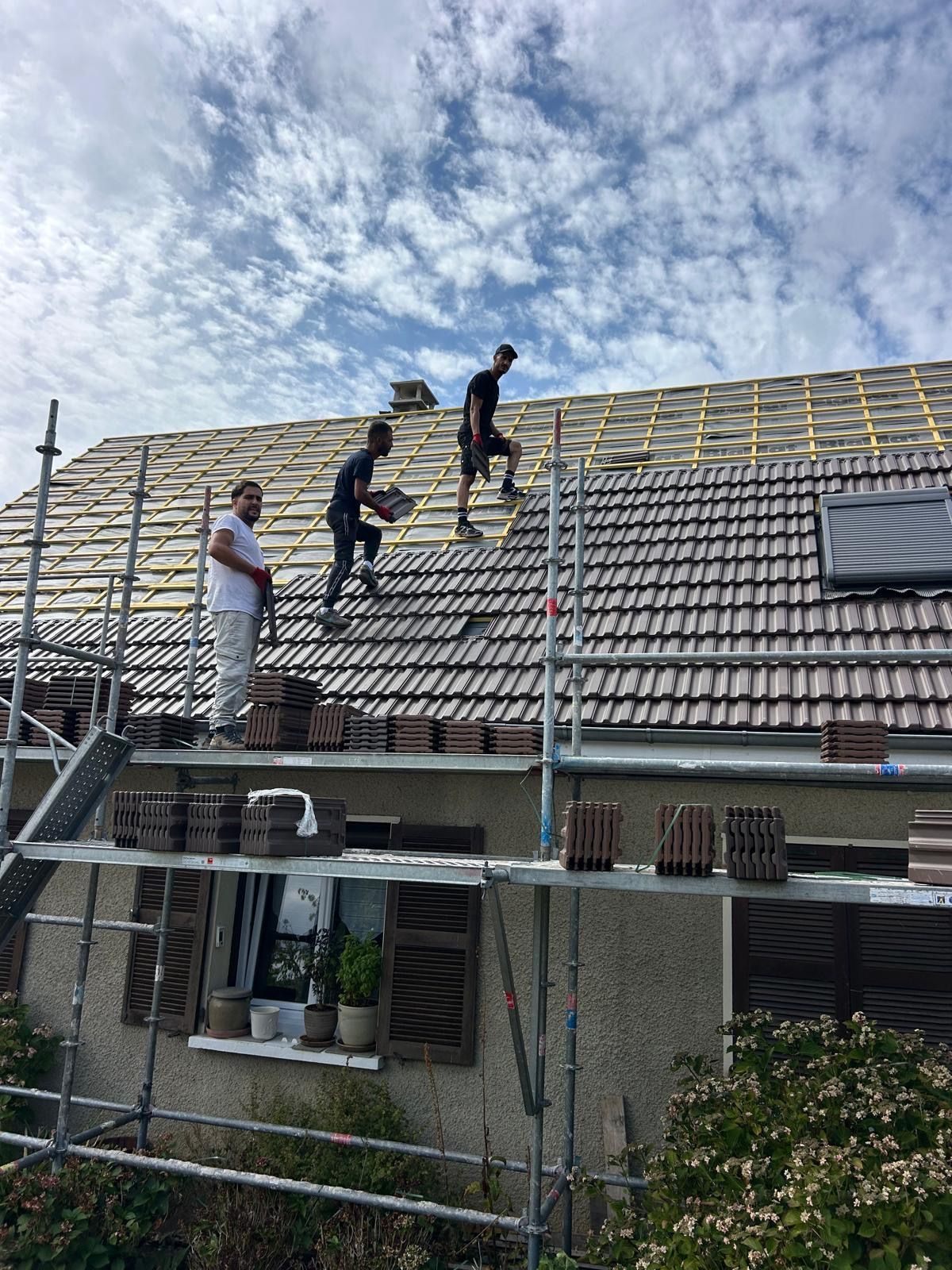 Des couvreurs travaillent sur le toit d'une maison en tuiles, des échafaudages sont présents tout autour de la maison.
