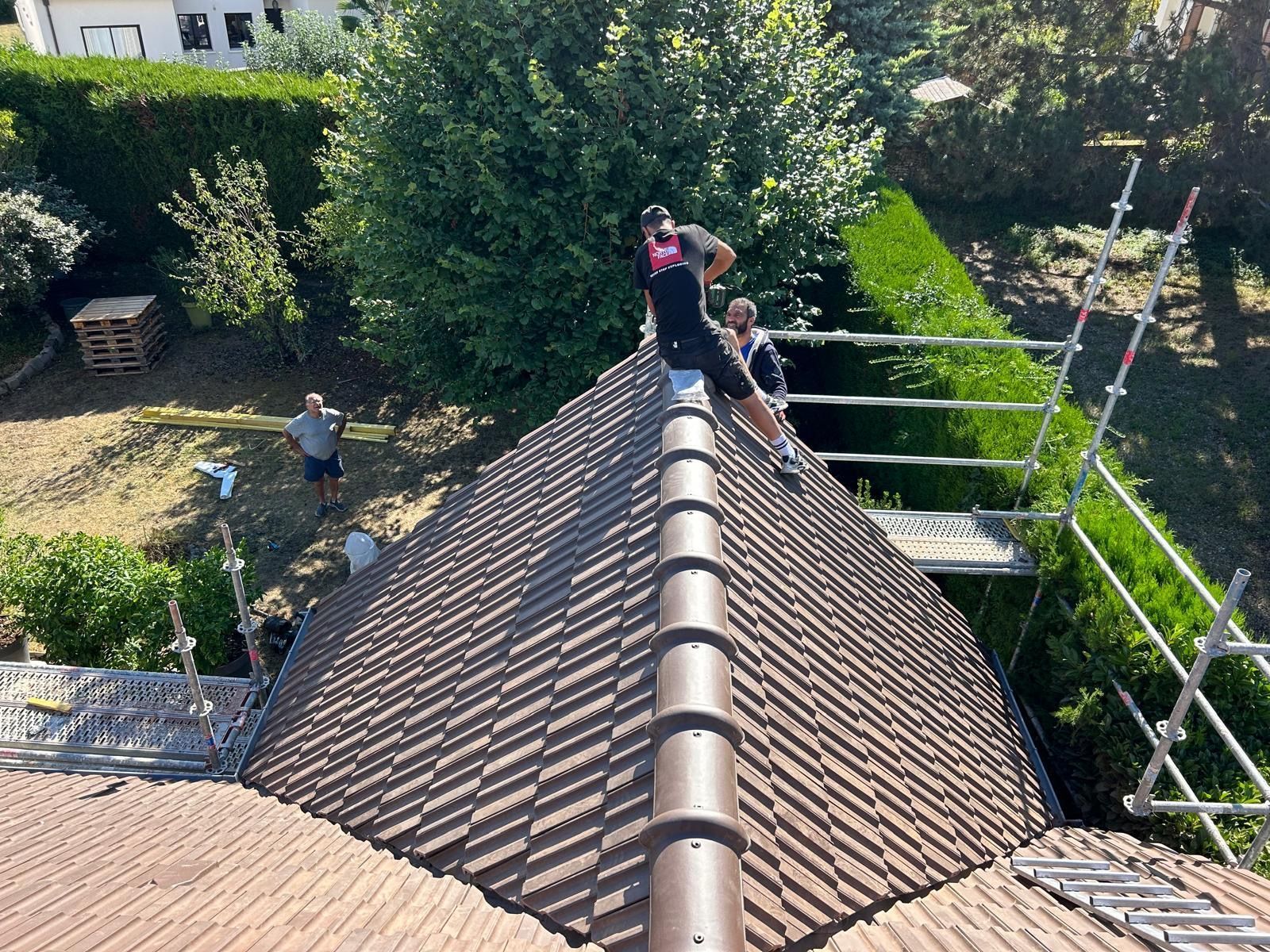 Deux ouvriers, équipés de harnais de sécurité, travaillent sur un toit de tuiles, la maison est entourée d'échafaudages.