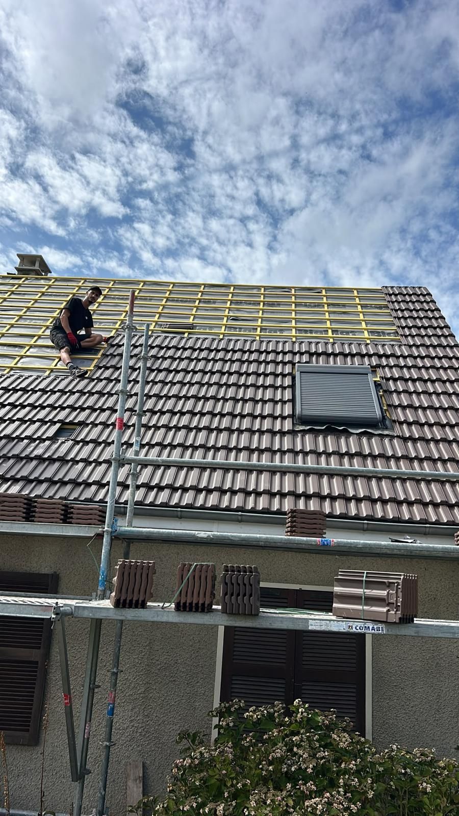 Un ouvrier installe de l'isolant jaune sur un toit de tuiles sous un ciel nuageux.