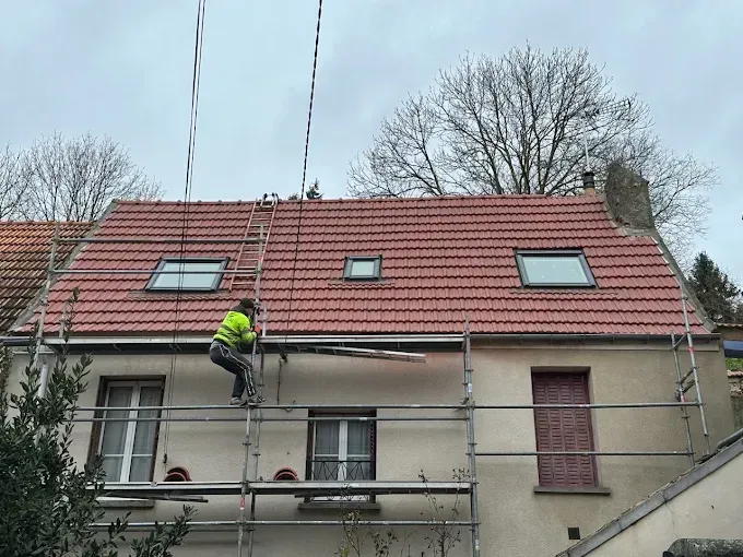 Un ouvrier sur un échafaudage répare le toit d'une maison avec des tuiles rouges et des fenêtres de toit.