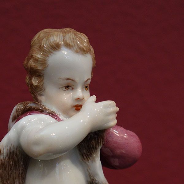 Engel aus Porzellan hält einen rosafarbenen Gegenstand vor einem burgunderfarbenen Hintergrund.