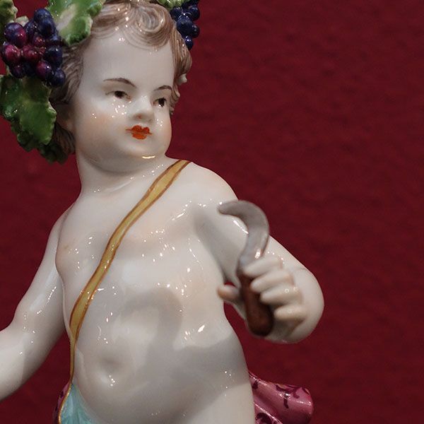 Porzellanengel mit Trauben, der eine Sense hält, vor rotem Hintergrund.
