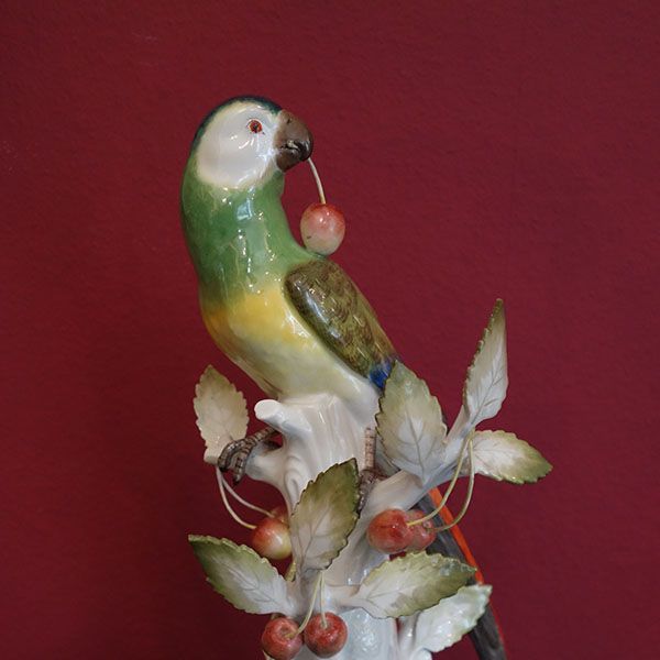Keramikpapagei auf einem Ast, der eine Kirsche hält. Grüne, gelbe und weiße Federn. Rote Kirschen und Hintergrund.