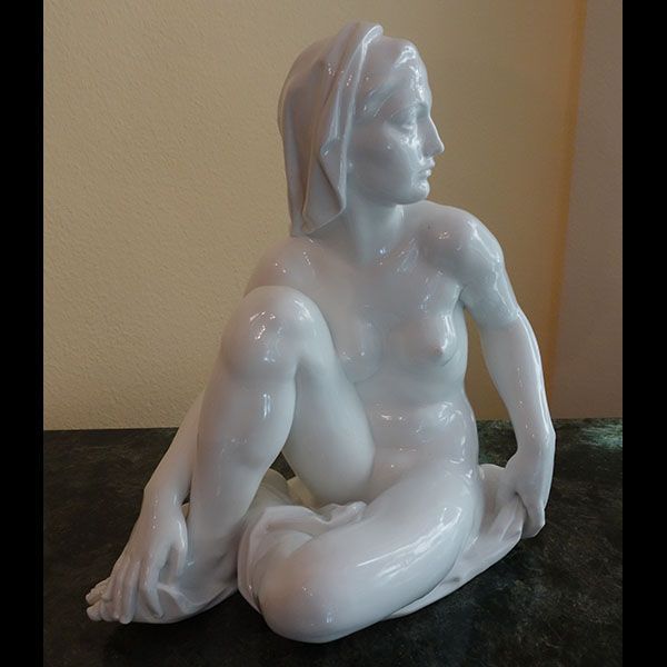 Weiße Porzellanskulptur einer sitzenden Frau mit drapiertem Kopf, die zur Seite blickt.