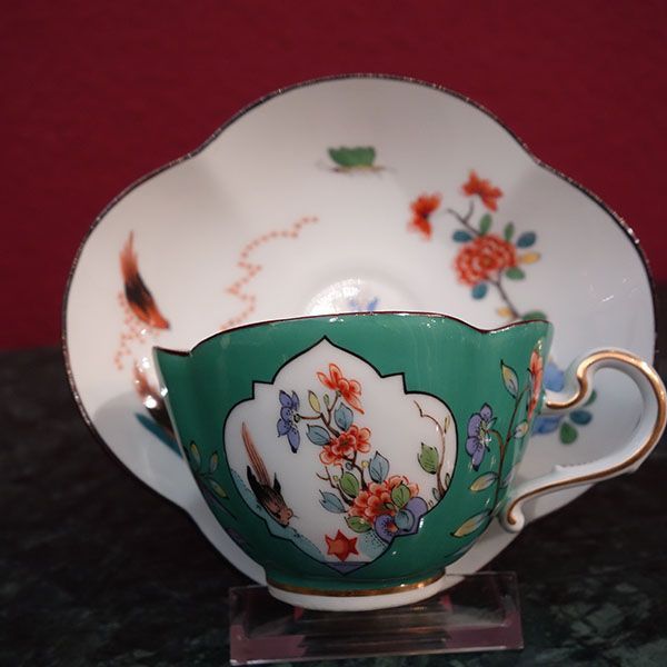 Grüne Teetasse und Untertasse mit Blumenmuster und Goldrand auf rotem Hintergrund.