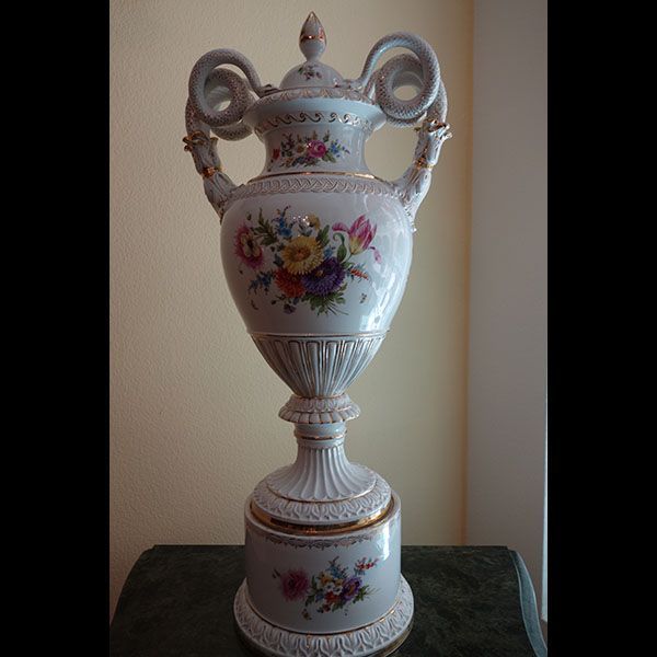 Hohe, kunstvolle Vase aus weißem Porzellan mit Deckel und Blumenmuster; auf einem Sockel und einem Tisch platziert.