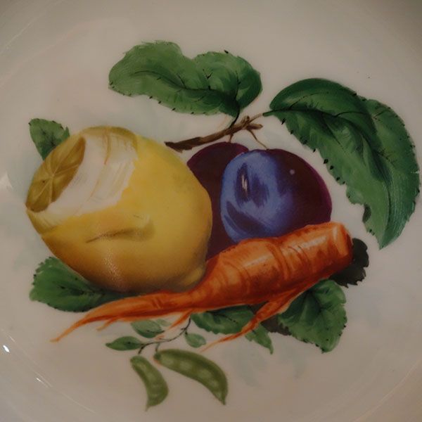 Teller mit handgemaltem Arrangement: gelbe Früchte, violette Pflaume, orangefarbenes Wurzelgemüse und grüne Blätter.