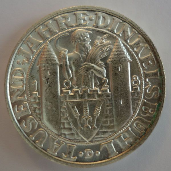 Eine Silbermünze mit einer zentralen Figur, die Weizen hält, flankiert von Türmen und umgeben von Text.