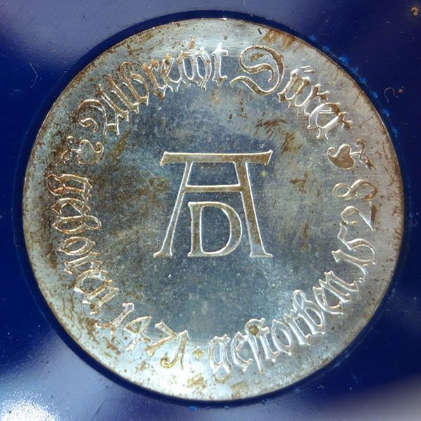 Silbernes rundes Medaillon mit erhabenem Text in einem Kreis um den Rand und „AD“ in der Mitte.