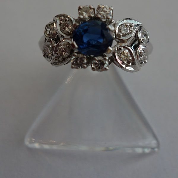 Ring mit einem großen blauen Stein, umgeben von kleineren Diamanten, präsentiert auf einem durchsichtigen Ständer.