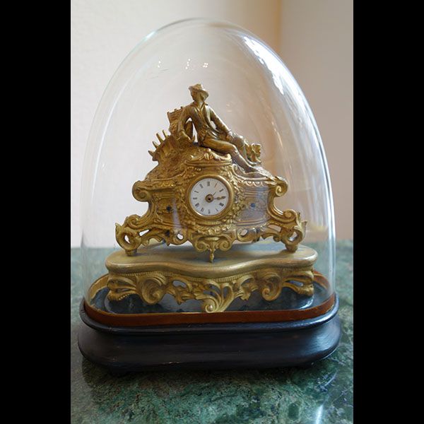 Verzierte goldene Uhr unter einer Glaskuppel auf einer Marmorplatte. Darauf liegt eine Figur.