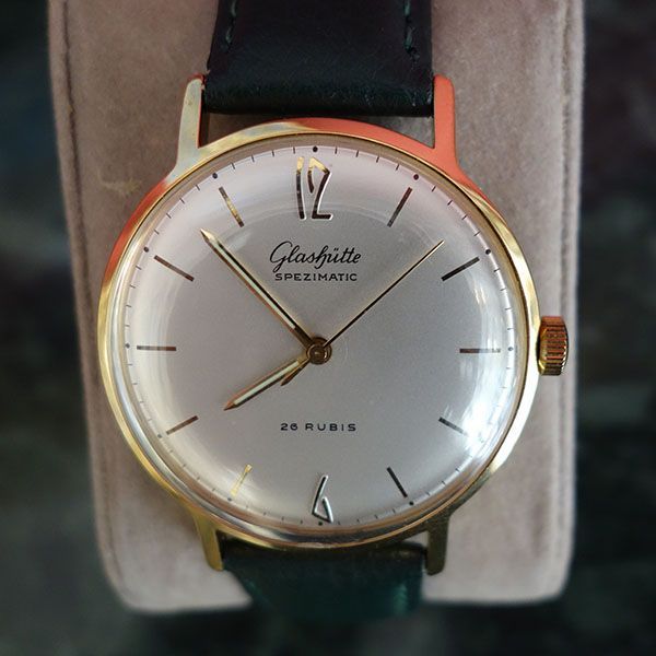 Goldene Glashütte Spezichron-Uhr mit silbernem Zifferblatt, goldenen Zeigern und Markierungen, schwarzem Armband.