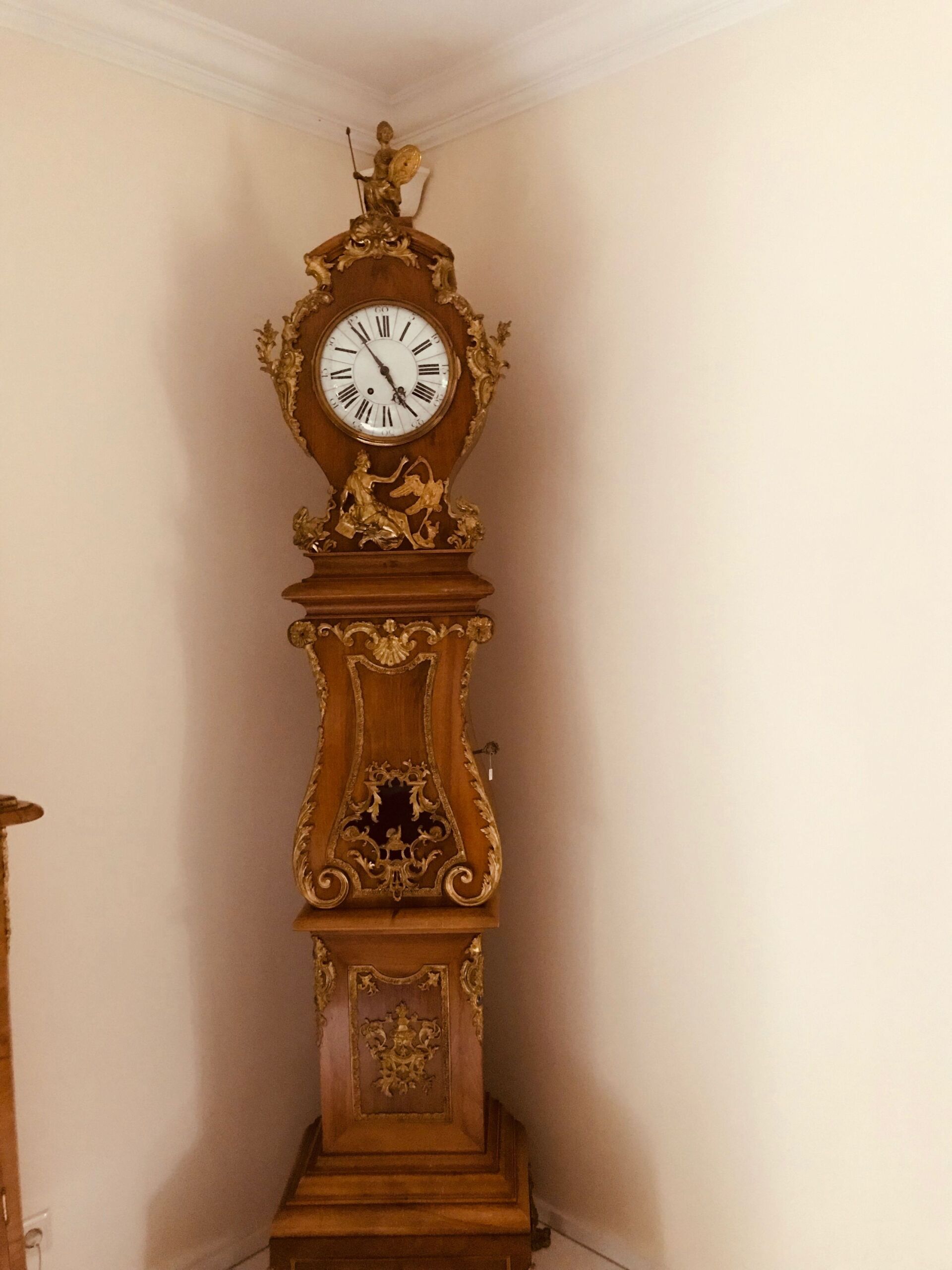 Verzierte antike Standuhr in einer Ecke vor beigen Wänden.