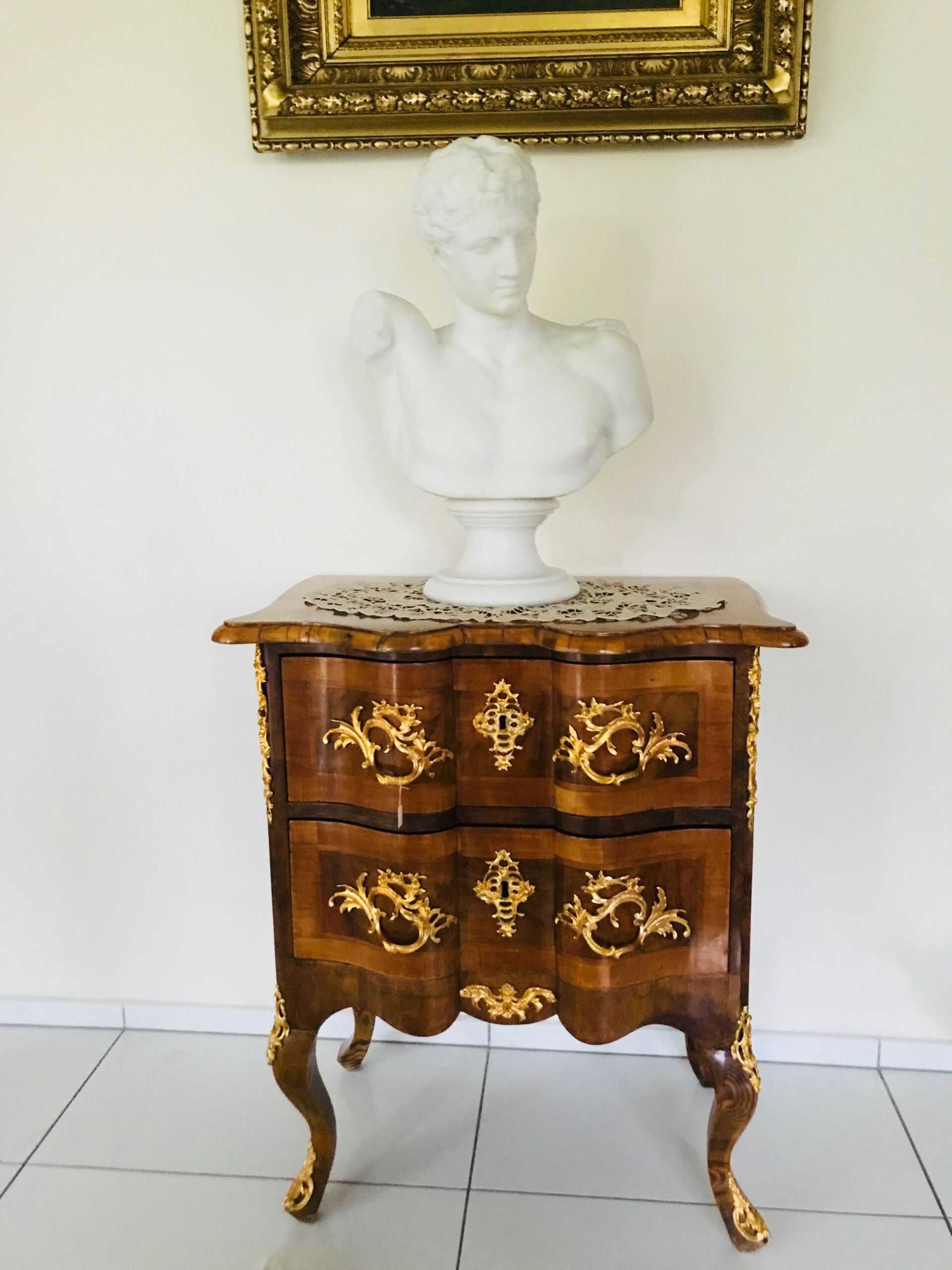 Eine Marmorbüste auf einem dekorativen Holzschrank mit goldenen Akzenten vor einer weißen Wand.