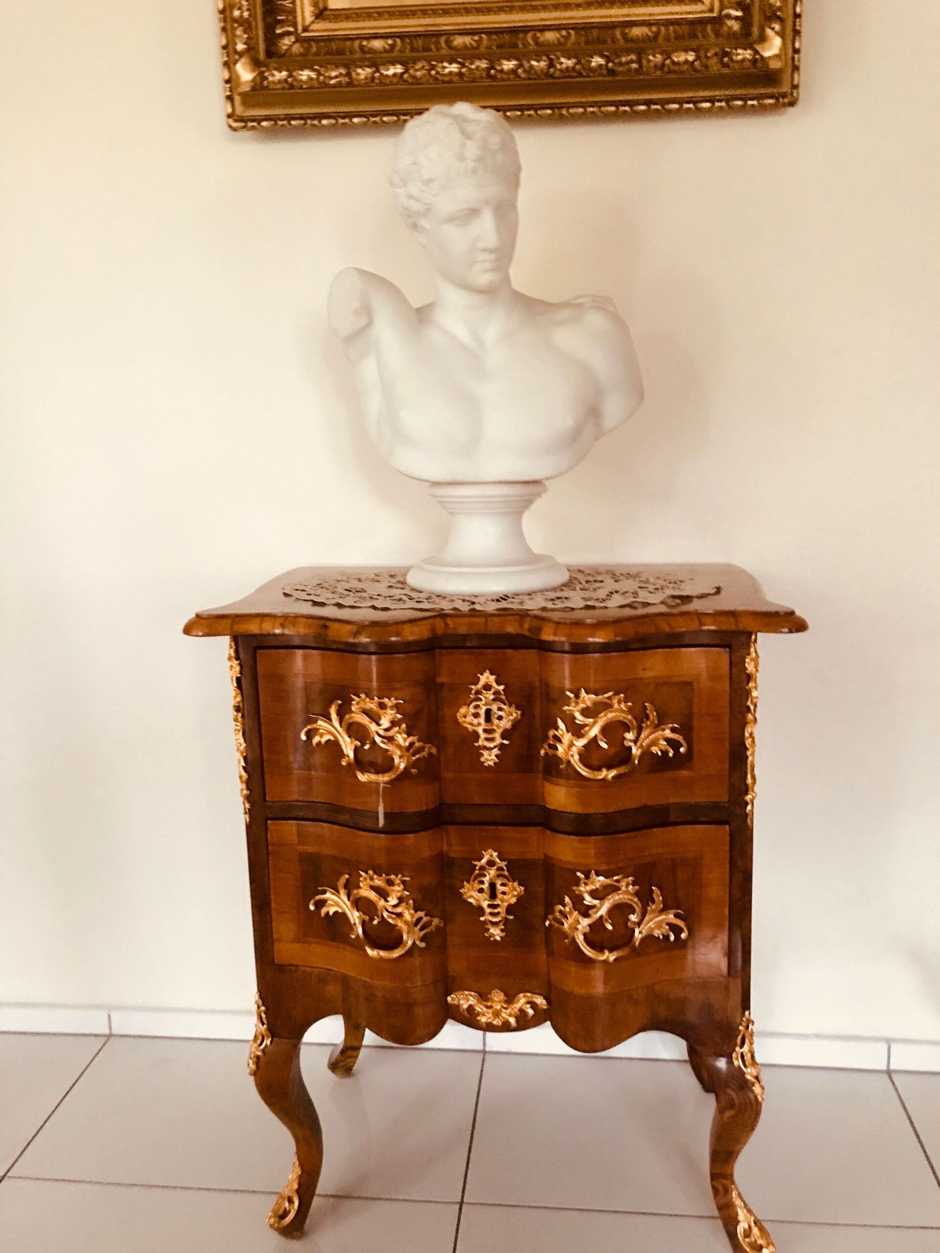 Marmorbüste auf verziertem Holzschrank, goldene Akzente, vor weißer Wand.