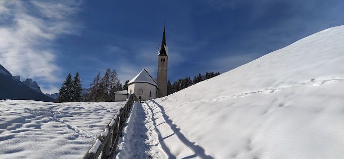 Un sentiero innevato conduce a una chiesa con un alto campanile, che si staglia contro un cielo azzurro brillante.