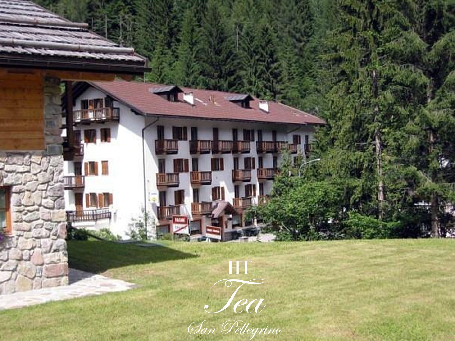 Edificio bianco a più piani con balconi e tetto marrone, con uno sfondo di alberi ed erba verde.