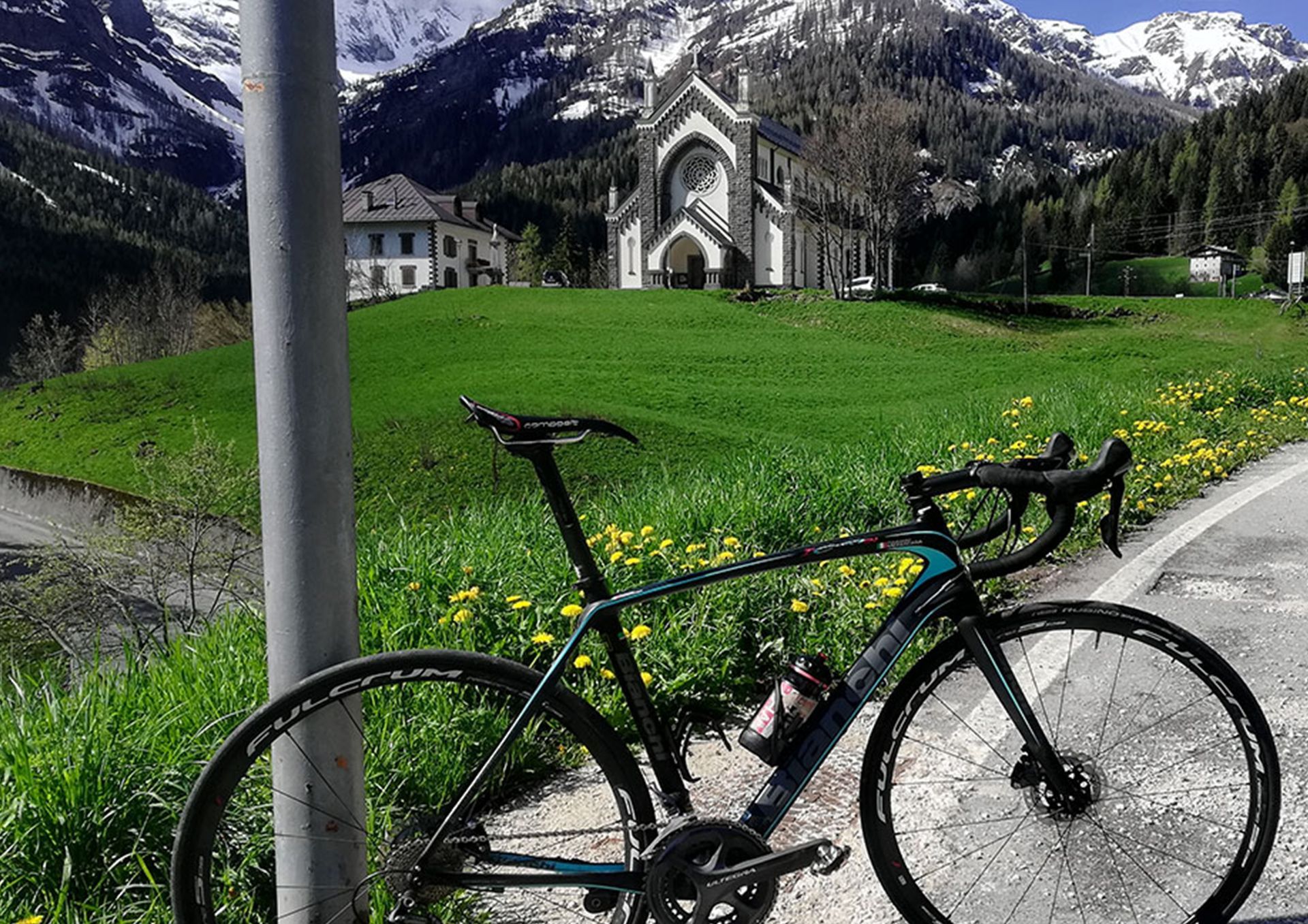 Bicicletta da strada parcheggiata vicino a un palo, con vista su una chiesa e sulle montagne in una lussureggiante valle verde.