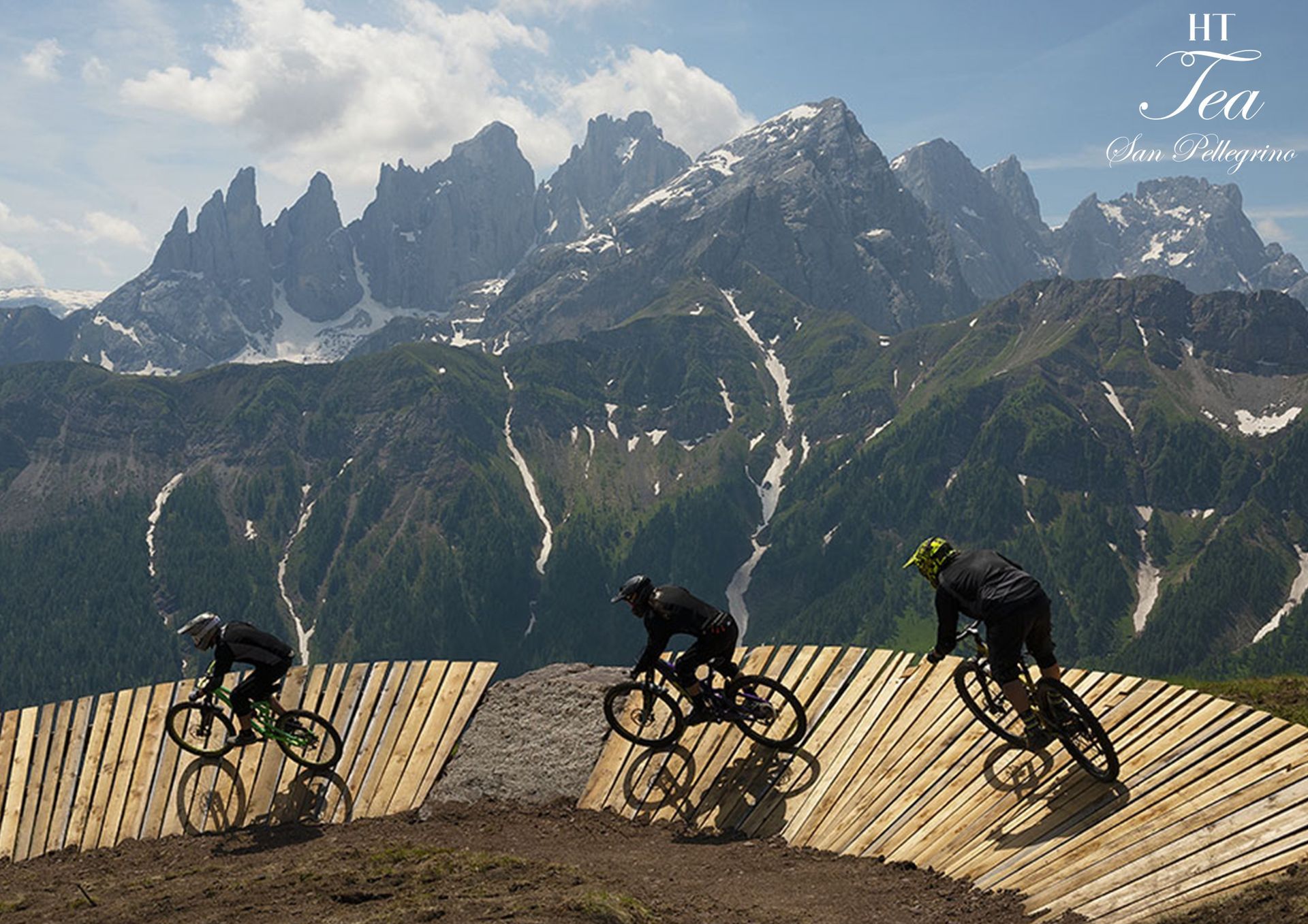 Tre ciclisti di montagna affrontano curve sopraelevate in legno su una pista sterrata con sullo sfondo montagne innevate.