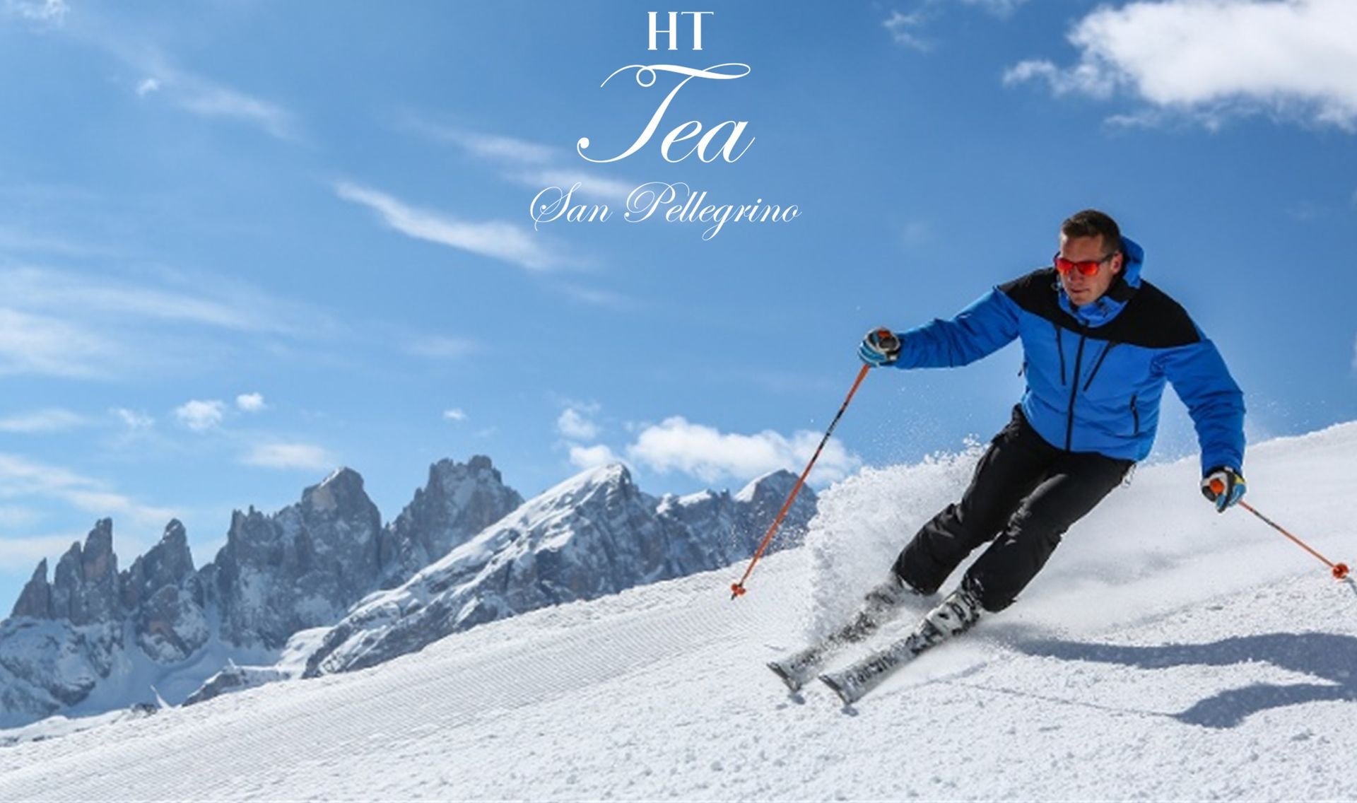 Uno sciatore con giacca blu scende lungo un pendio innevato con montagne e cielo azzurro sullo sfondo; logo IIT Tea.