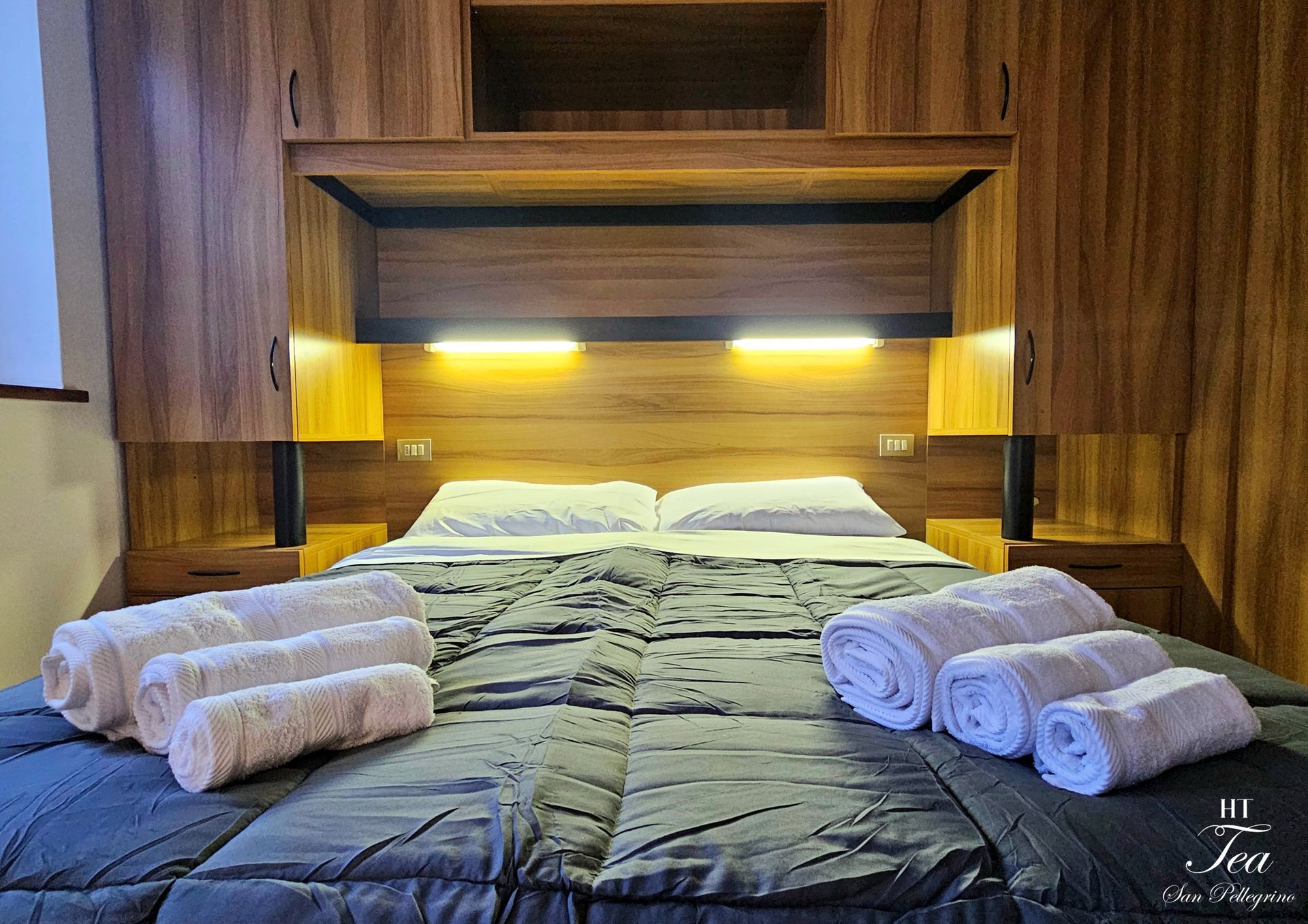 Camera da letto con testiera in legno, contenitore sopraelevato, letto con lenzuola grigie e asciugamani arrotolati.