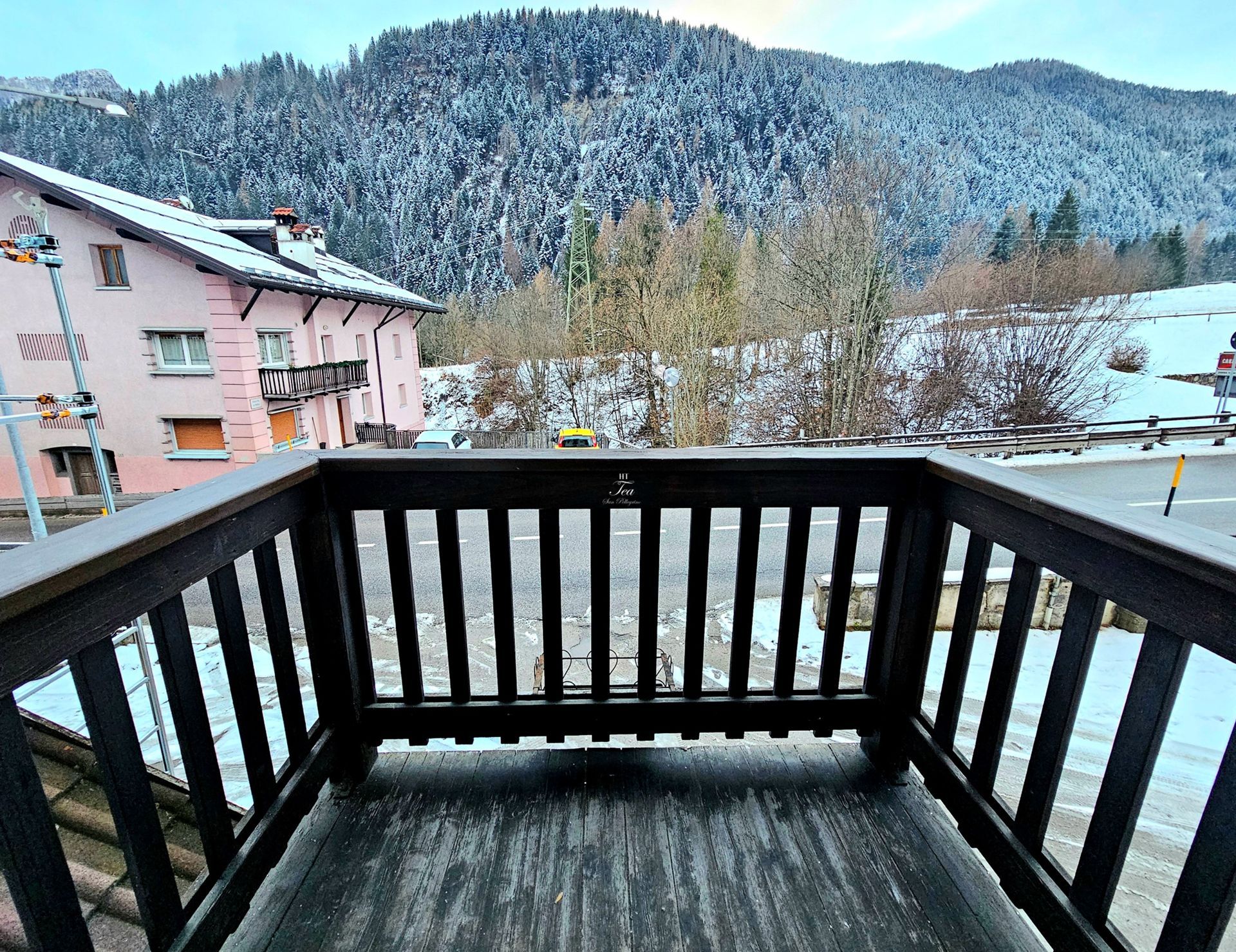 Balcone in legno con vista su una montagna innevata e su un villaggio. Sulla sinistra si trova un edificio rosa.