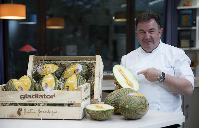 Chef señalando un melón cortado por la mitad, con una caja de melones frente a él. Dentro de una cocina.
