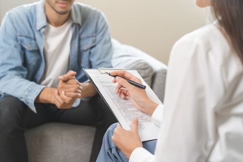 Un hombre está sentado en un sofá hablando con una mujer que sostiene un portapapeles.