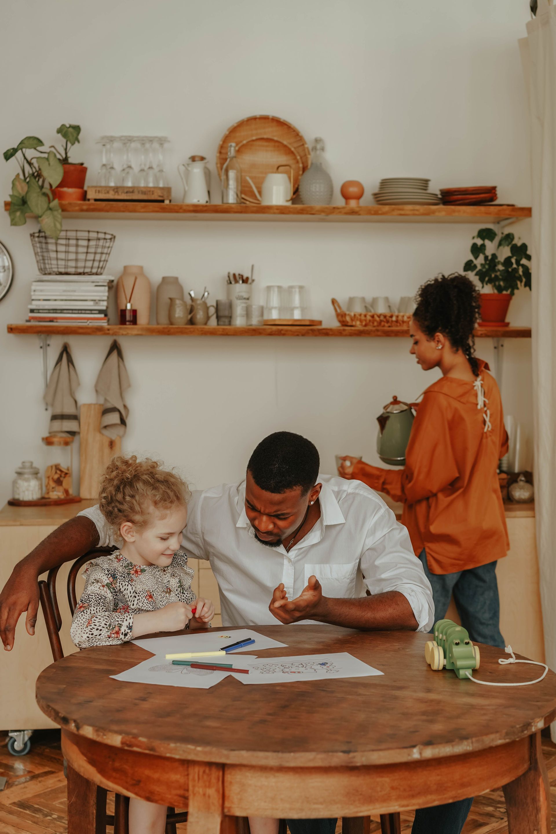 Trois personnes réunies autour d'une table en bois, travaillant sur des documents dans une cuisine lumineuse avec des étagères et des plantes.