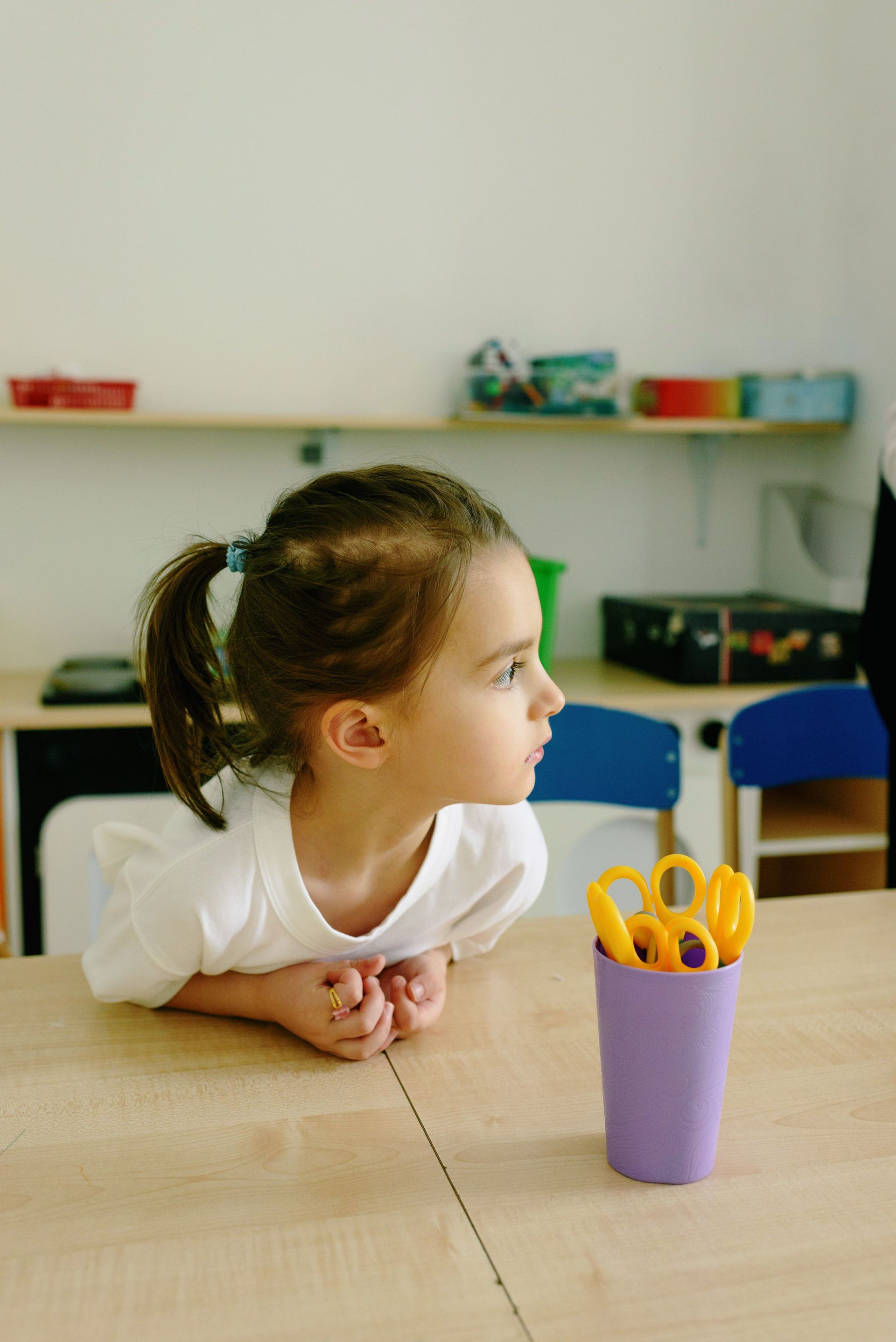Enfant appuyé sur une table à côté d'un gobelet violet contenant des ciseaux dans une salle de classe