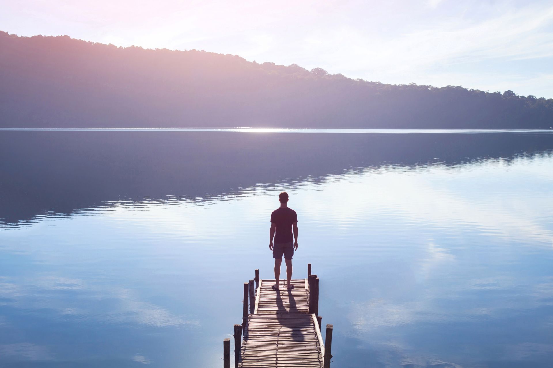 Personne debout sur un quai en bois, face à un lac calme reflétant le lever du soleil, avec une colline boisée en arrière-plan.